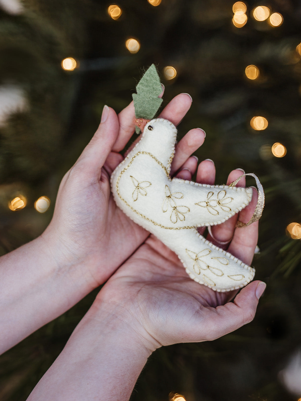 Artisan-crafted white felt dove ornament with gold embroidery held in hands, Christmas tree lights softly glowing in the background