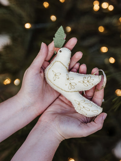 Artisan-crafted white felt dove ornament with gold embroidery held in hands, Christmas tree lights softly glowing in the background