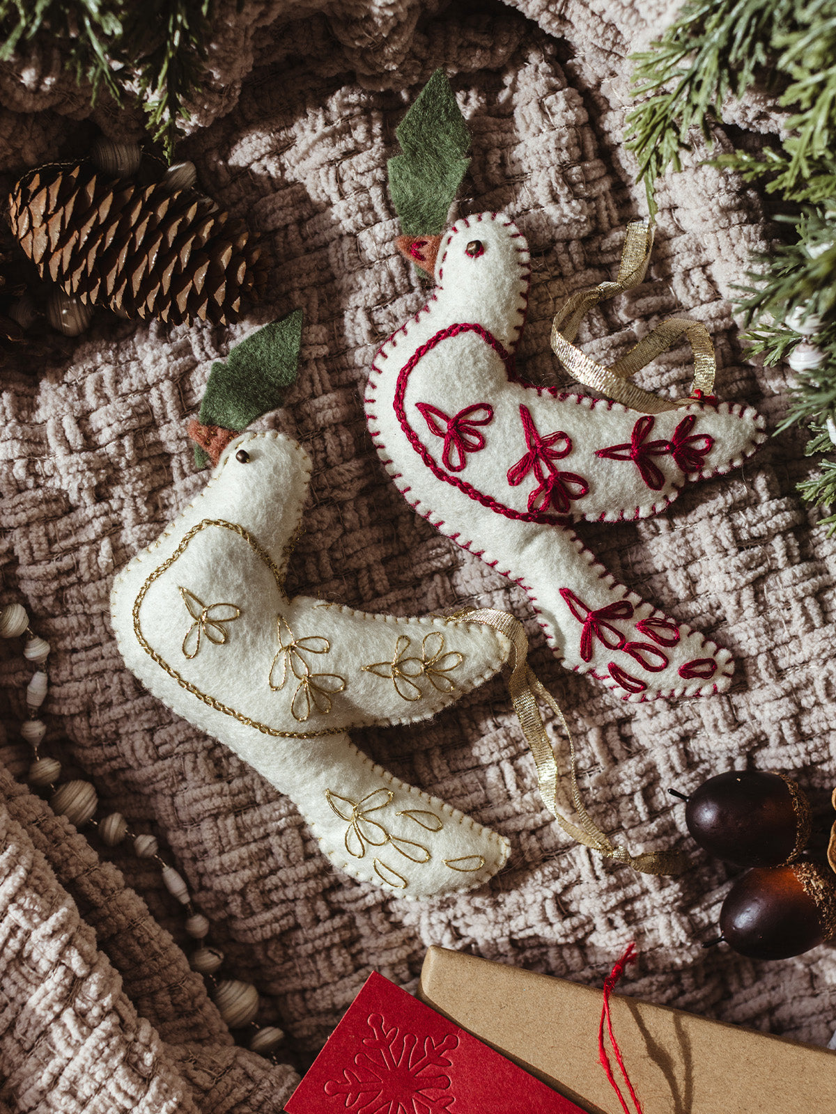 Set of two artisan felt dove ornaments, one with red stitching and one with gold embroidery, displayed on cozy textured blanket with pinecones – sustainable Christmas decorations