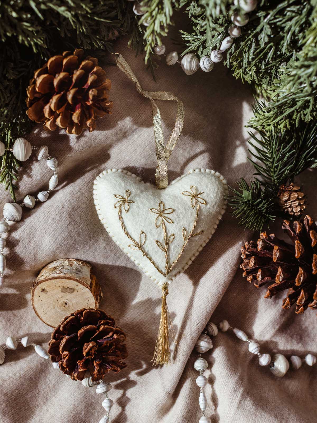 Handcrafted white felt heart ornament with gold embroidery and tassel, styled with pinecones, evergreen branches, and neutral linens — a cozy, artisan holiday decoration.