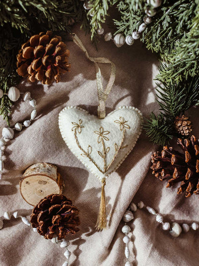 Handcrafted white felt heart ornament with gold embroidery and tassel, styled with pinecones, evergreen branches, and neutral linens — a cozy, artisan holiday decoration.