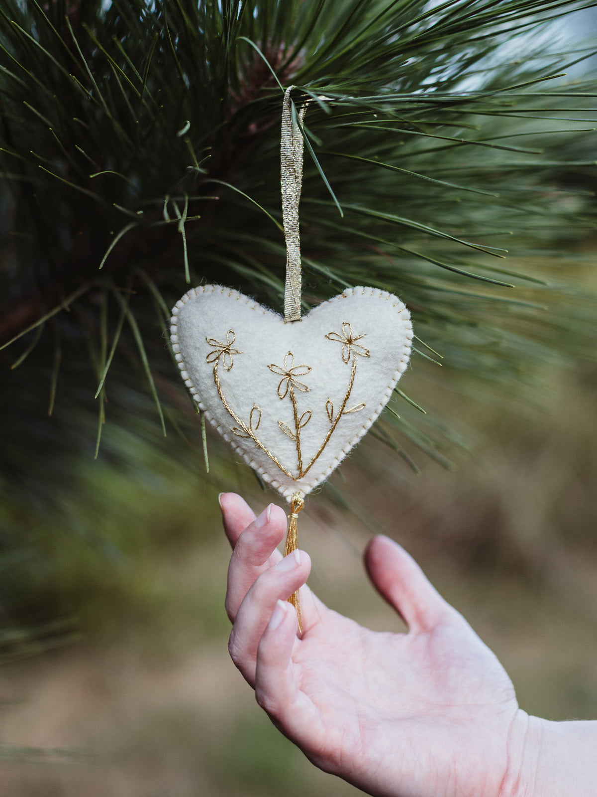 Hand holding a white felt heart ornament with gold floral embroidery and tassel, hanging from a pine branch — highlighting the handmade craftsmanship and festive detail.