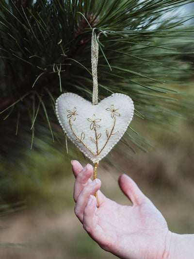 Hand holding a white felt heart ornament with gold floral embroidery and tassel, hanging from a pine branch — highlighting the handmade craftsmanship and festive detail.