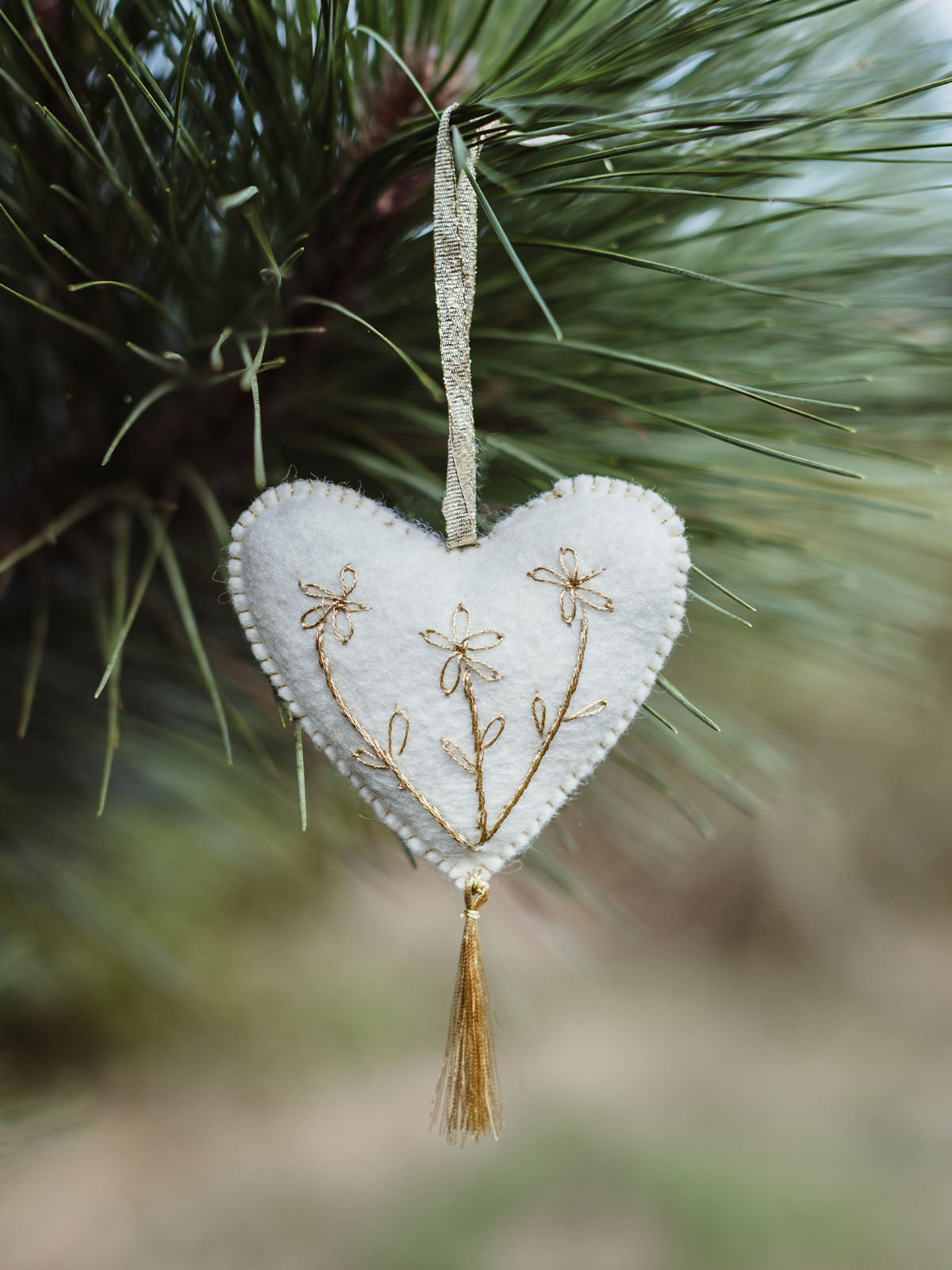 White felt heart ornament with delicate gold floral embroidery and tassel, hanging on a pine tree branch for a simple, natural Christmas decor look.