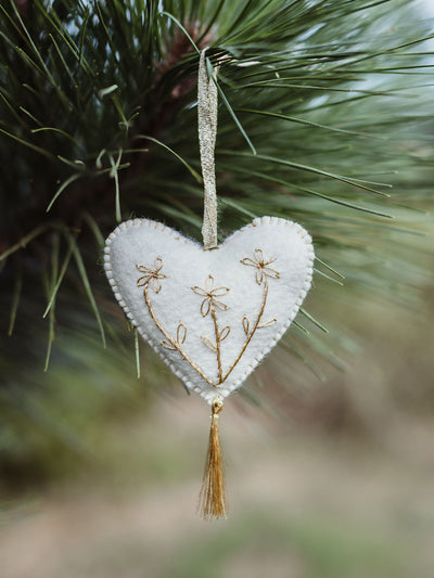 White felt heart ornament with delicate gold floral embroidery and tassel, hanging on a pine tree branch for a simple, natural Christmas decor look.