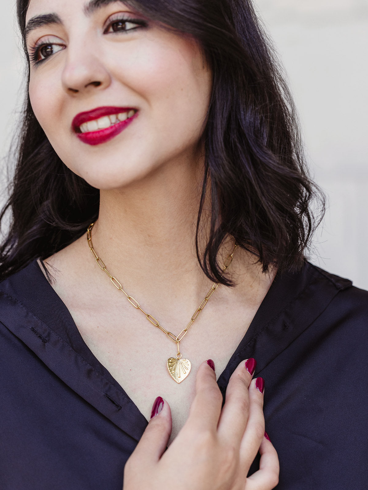 Woman with dark hair and red lipstick wearing a gold chain necklace with a heart-shaped leaf pendant, styled with a black blouse and red manicure.