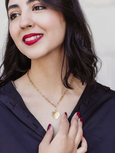 Woman with dark hair and red lipstick wearing a gold chain necklace with a heart-shaped leaf pendant, styled with a black blouse and red manicure.