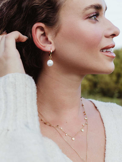 Woman in cozy cream sweater wearing layered gold coin necklaces and gold hoop earrings with a freshwater pearl drop – sustainable jewelry style.