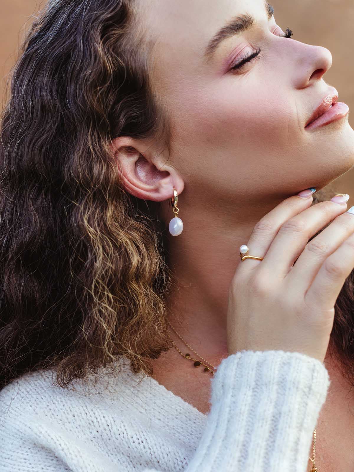 Woman with wavy hair wearing gold pearl drop hoop earrings and matching pearl ring, styled with soft knit sweater – artisan-made jewelry.