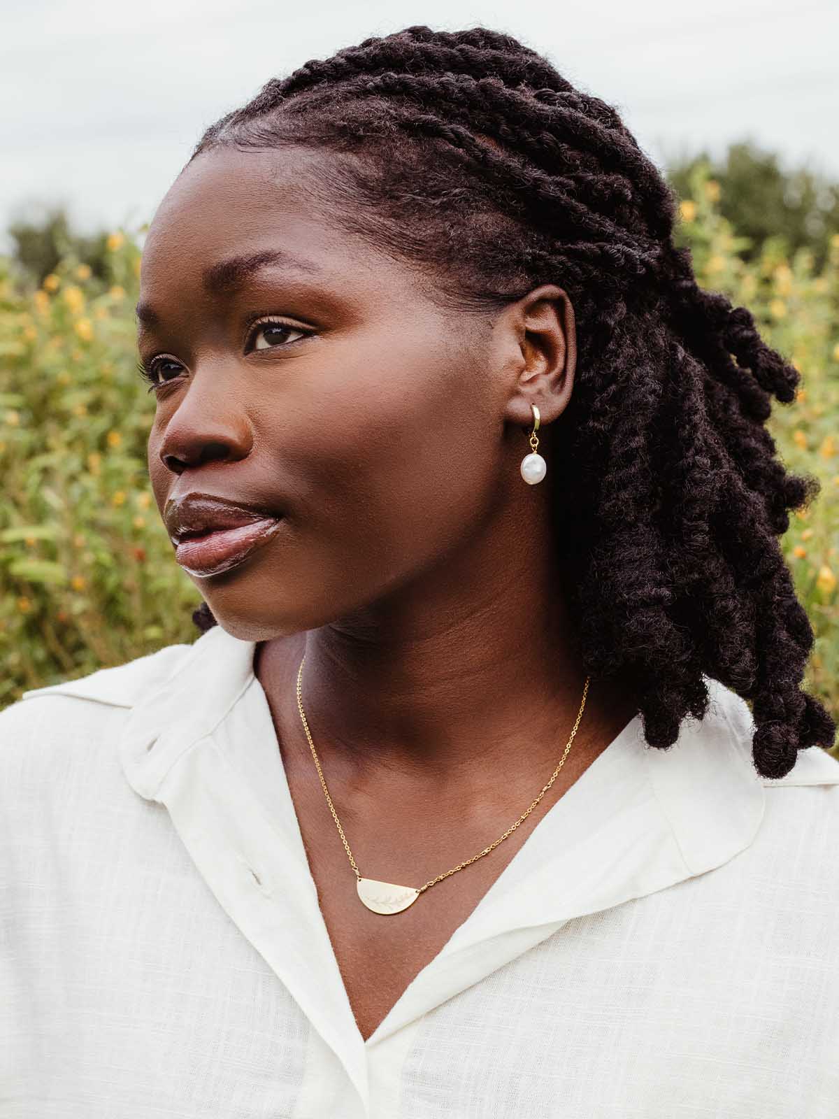 Woman outdoors in white button-up top wearing gold hoop earrings with pearl drop and half-moon gold pendant necklace – ethically made jewelry.