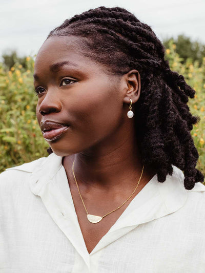 Woman outdoors in white button-up top wearing gold hoop earrings with pearl drop and half-moon gold pendant necklace – ethically made jewelry.