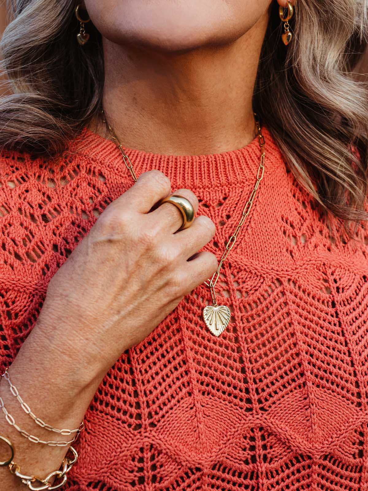 Detail shot of woman’s hand holding a gold heart pendant necklace, styled with gold bracelet and coral knit sweater.