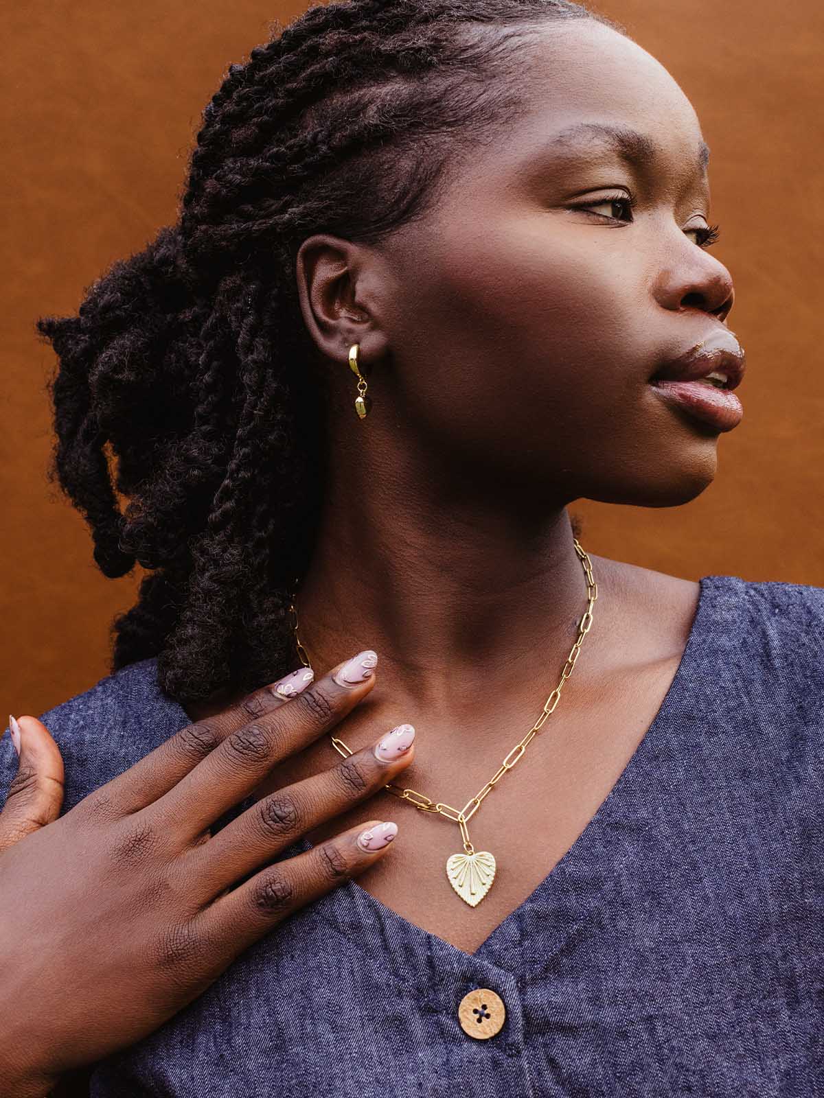 Close-up of woman in denim dress wearing a gold chain necklace with a heart-shaped leaf pendant and gold dangle earrings, highlighting ethical handmade jewelry.