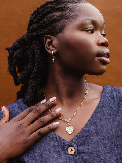 Close-up of woman in denim dress wearing a gold chain necklace with a heart-shaped leaf pendant and gold dangle earrings, highlighting ethical handmade jewelry.