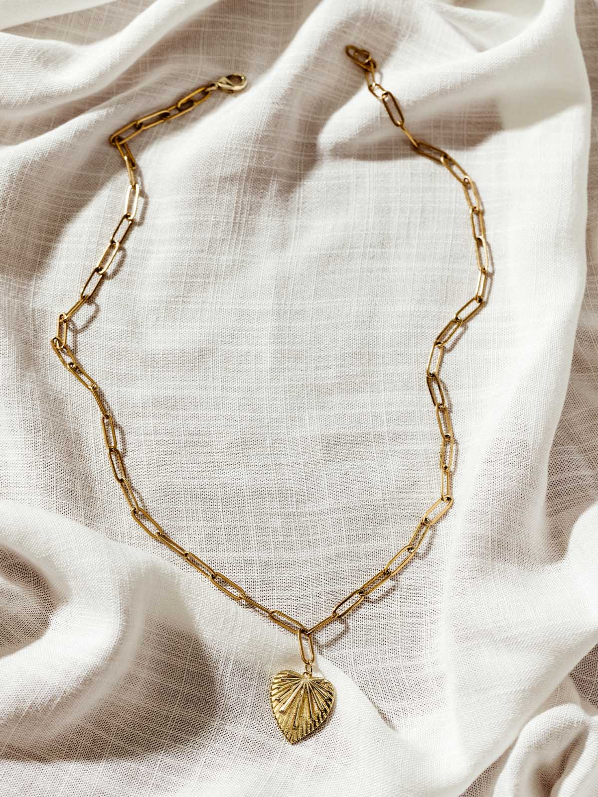 Gold chain link necklace with textured heart pendant displayed on neutral linen fabric – ethical and stylish accessory for layering.