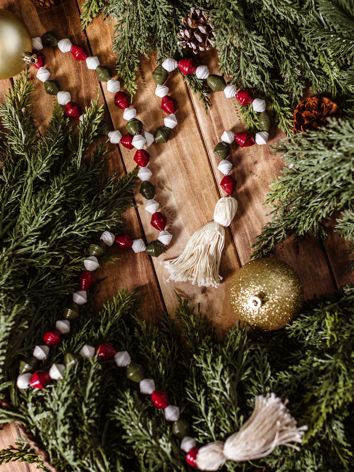 Ethically made holiday garland featuring red, white, and green paper beads with tassel ends, styled with evergreen branches and gold ornament.