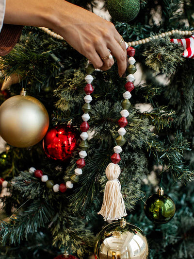 Person decorating a Christmas tree with a sustainable red, green, and white paper bead garland featuring tassels among gold and red ornaments.