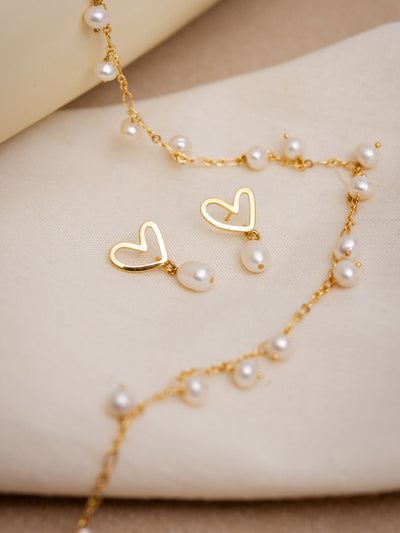 Delicate gold heart drop earrings with freshwater pearl charms styled on a soft neutral fabric with a dainty gold and pearl chain. Handmade ethical jewelry perfect for gifting or bridal accessories.