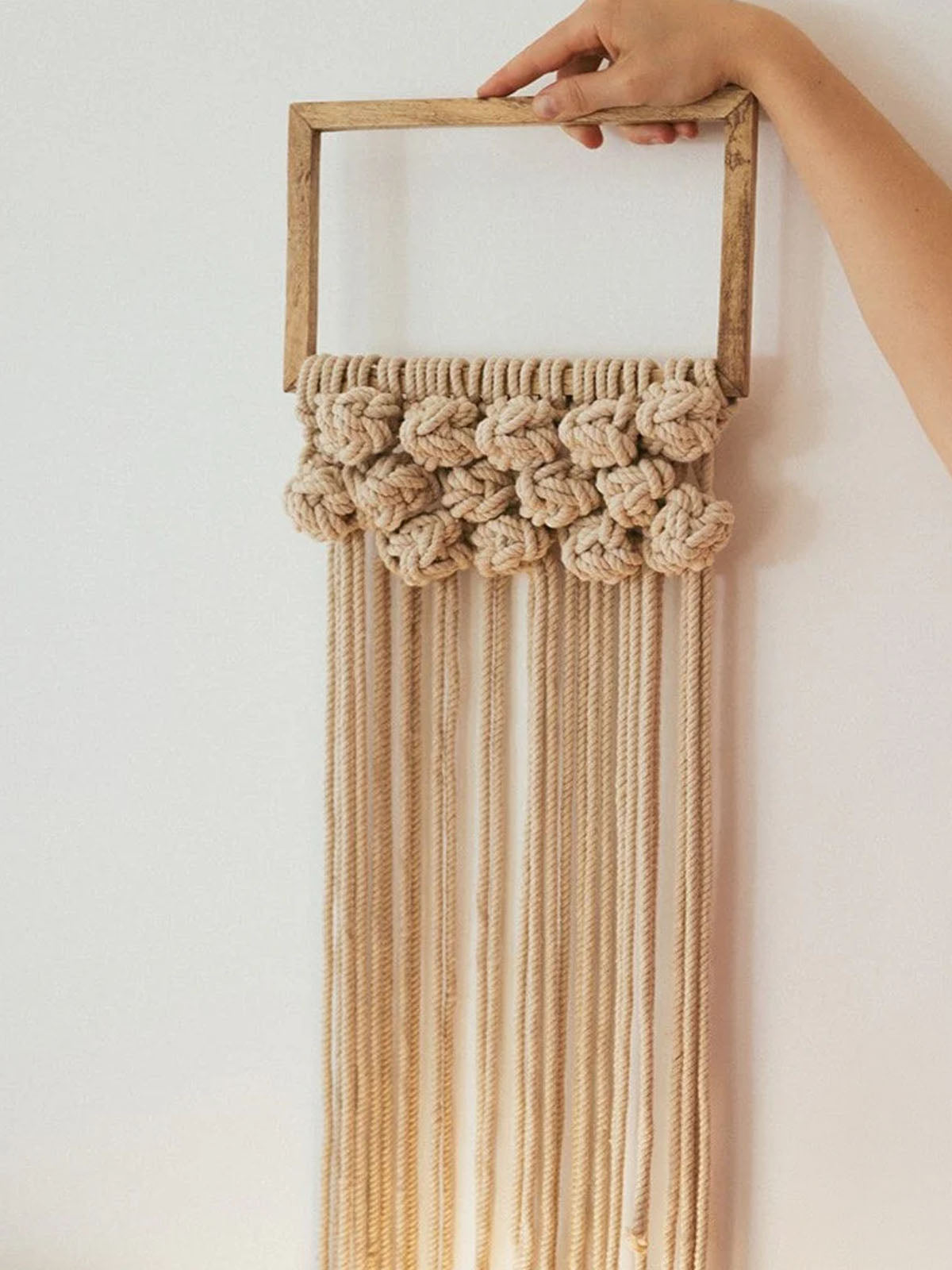 Hand holding rope wall hanging over white wall with square wooden top that has wall hanging hardware. 