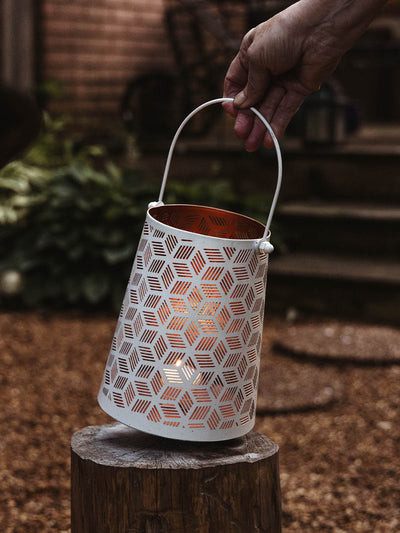 A hand holding a white metal lantern with a geometric cutout pattern, lit from inside and resting on a tree stump in a garden at dusk.