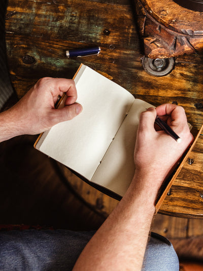 Overhead view of a person writing in a leather journal with blank cream-colored pages, seated at a rustic wooden table with a pen cap nearby.