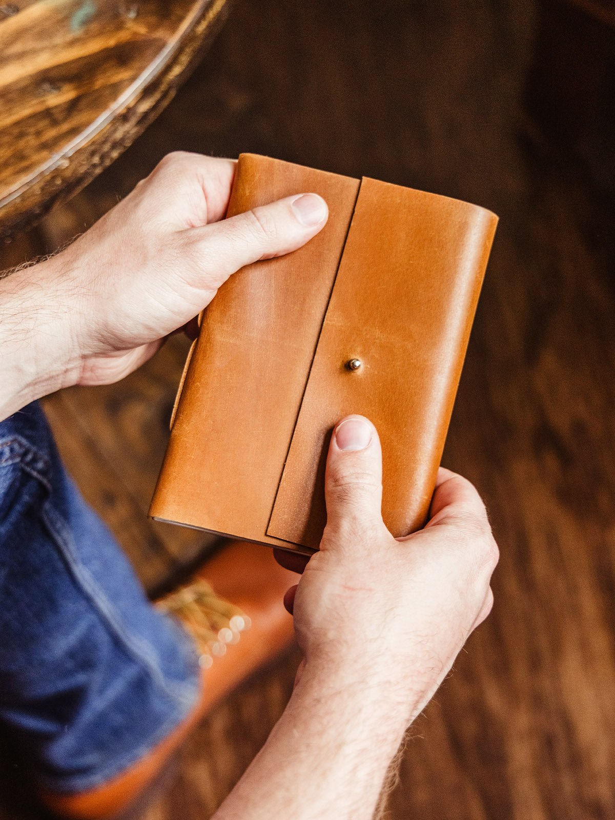 Hands holding a minimalist leather journal with a smooth tan finish and a brass button closure, over dark wood flooring.