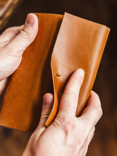 Close-up of hands opening a tan leather journal secured with a brass button, showcasing the supple texture of the cover.