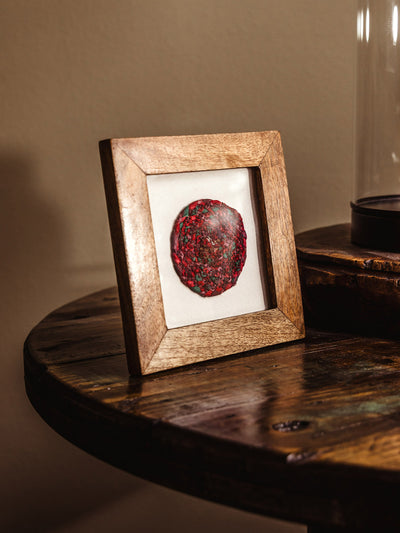 Framed red and green circular paper mosaic displayed on a wooden table next to a rustic carved object and glass dome.