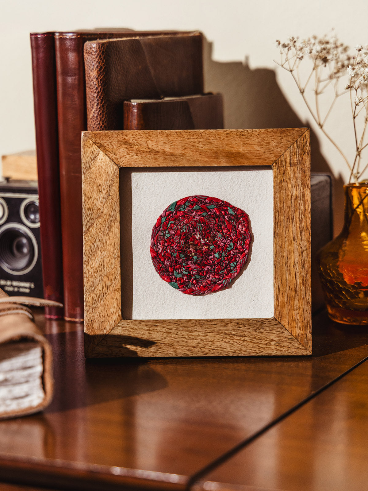 Framed artisan-made red circular mosaic resting on a wooden surface, styled with vintage books, dried florals, and a leather journal in the background.