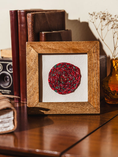 Framed artisan-made red circular mosaic resting on a wooden surface, styled with vintage books, dried florals, and a leather journal in the background.