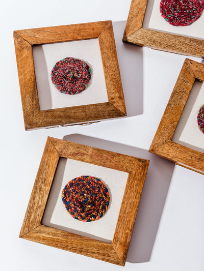 Handcrafted circular textile art piece in a wooden shadow box frame, featuring a dark multicolored woven design on a neutral background.