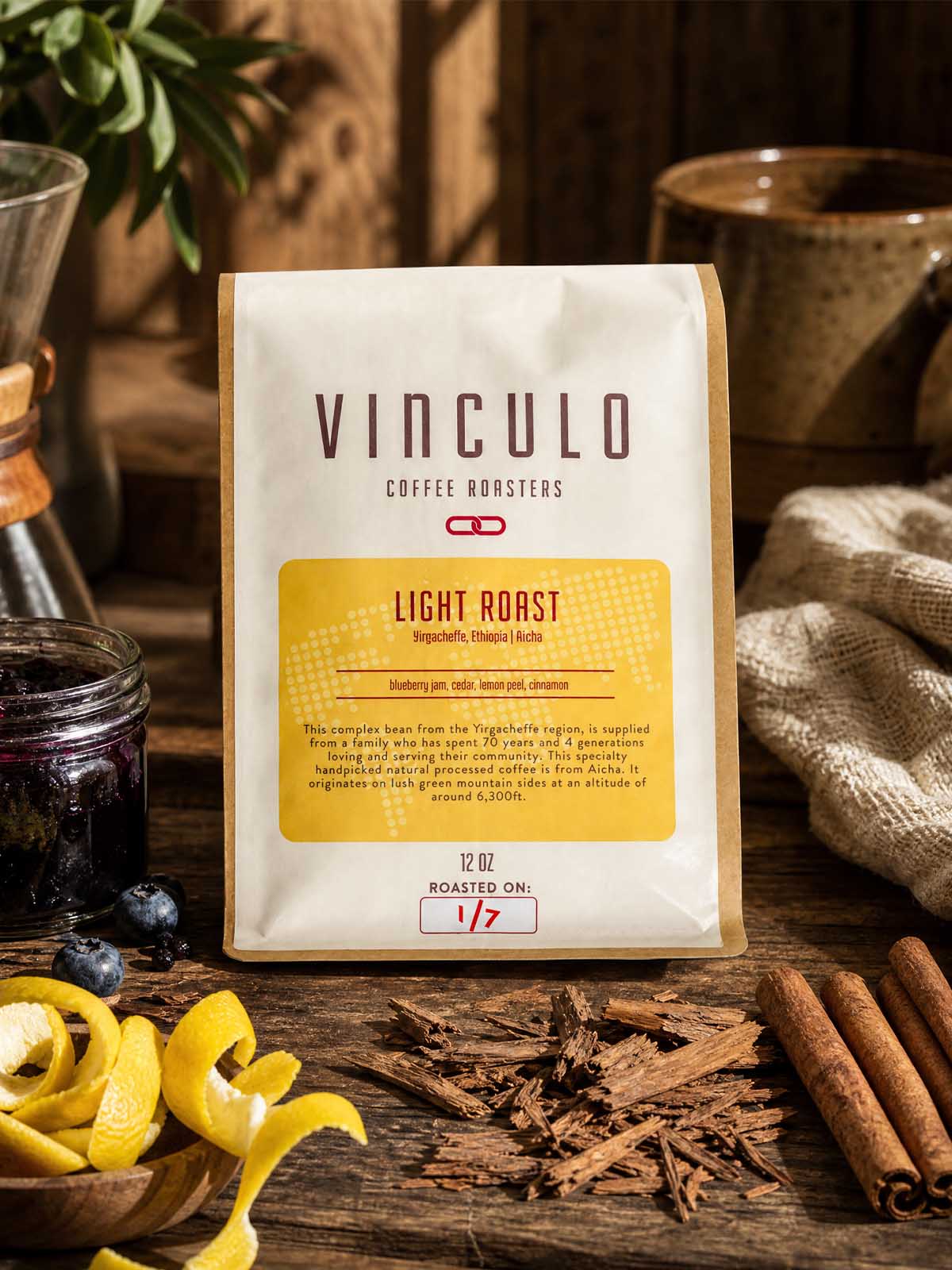 A bag of Vinculo Coffee Roasters Light Roast coffee stands on a rustic wooden table surrounded by coffee-themed props including cinnamon sticks, blueberries, lemon zest, a cloth, and ceramic mugs.