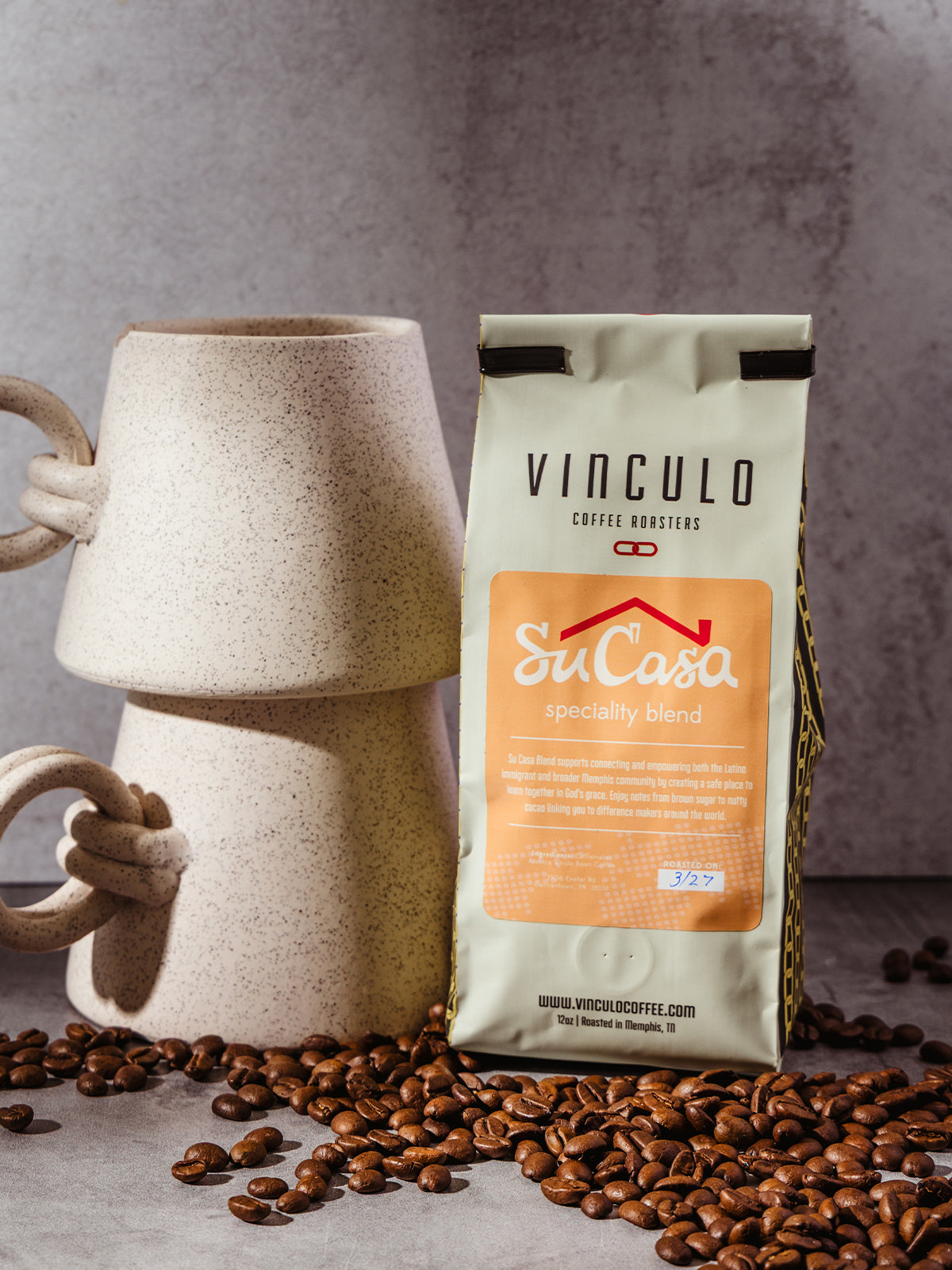 Vinculo Coffee Roasters Su Casa specialty blend bag placed beside a stacked ceramic mug, surrounded by scattered roasted coffee beans on a light stone surface.