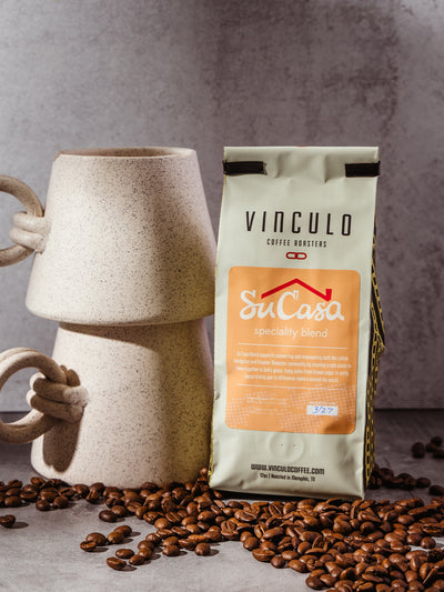 Vinculo Coffee Roasters Su Casa specialty blend bag placed beside a stacked ceramic mug, surrounded by scattered roasted coffee beans on a light stone surface.