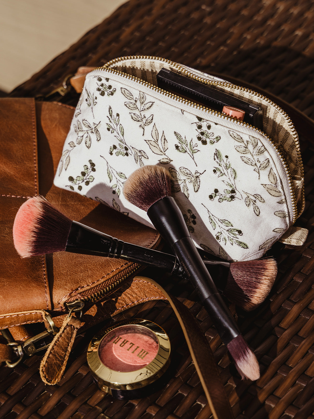 A makeup pouch with a leafy floral print rests beside a leather bag, surrounded by makeup brushes and blush. The pouch is unzipped, revealing neatly organized beauty products inside.