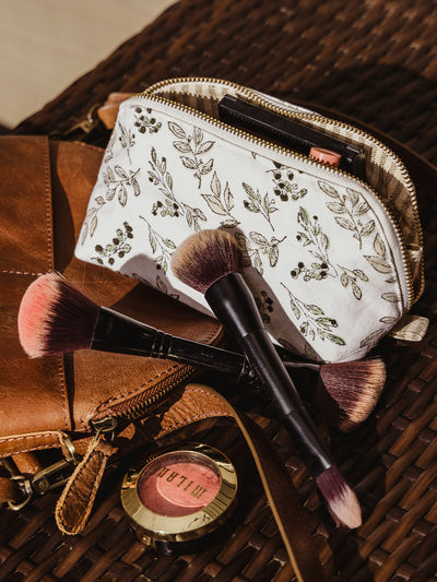 A makeup pouch with a leafy floral print rests beside a leather bag, surrounded by makeup brushes and blush. The pouch is unzipped, revealing neatly organized beauty products inside.