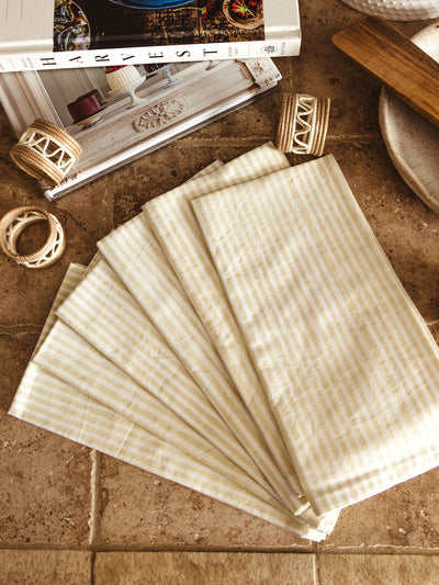 A set of neatly fanned beige and white striped cloth napkins displayed on a stone countertop, surrounded by handcrafted woven napkin rings in neutral tones and an open cookbook. Wooden utensils and ceramic dishware add to the cozy kitchen ambiance.