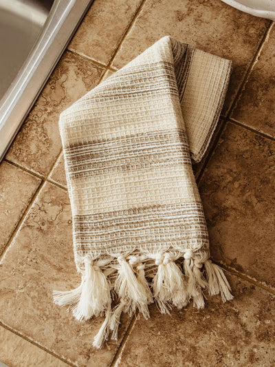 Neutral-toned striped kitchen towel with tasseled edges draped over a farmhouse sink. The setting features a stainless steel faucet, tiled backsplash, and neatly arranged dishwashing accessories.