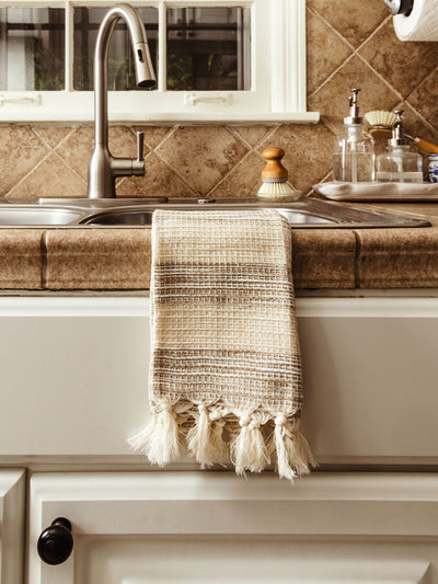 A beige and cream striped hand towel with a textured waffle weave and knotted tassels, neatly folded and resting on a tiled kitchen countertop near a sink.