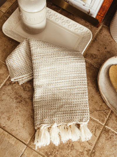 A cream and taupe waffle-weave kitchen towel with knotted fringe edges, folded on a tiled countertop beside a ceramic salt and pepper set and decorative dishware.