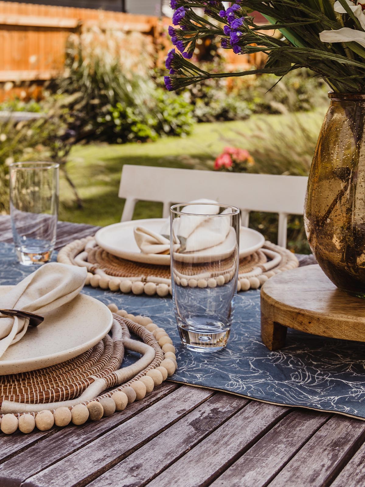 Place settings with plates and glasses on hibiscus table runner on a wooden table outside.