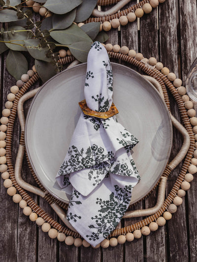 White napkin with a floral jade embroidery place on top of a plate.  Napkin, plate, and charger complete a full table setting on a wooden table with greenery.