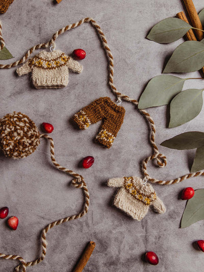 Handmade sweater garland with tan sweaters and brown pants hung on garland with little white wire hangers. Garland string is brown and tan with brown and cream pom poms on each end of the garland. Garland is seen on a concrete counter with cranberries, cinnamon sticks, and greenery.