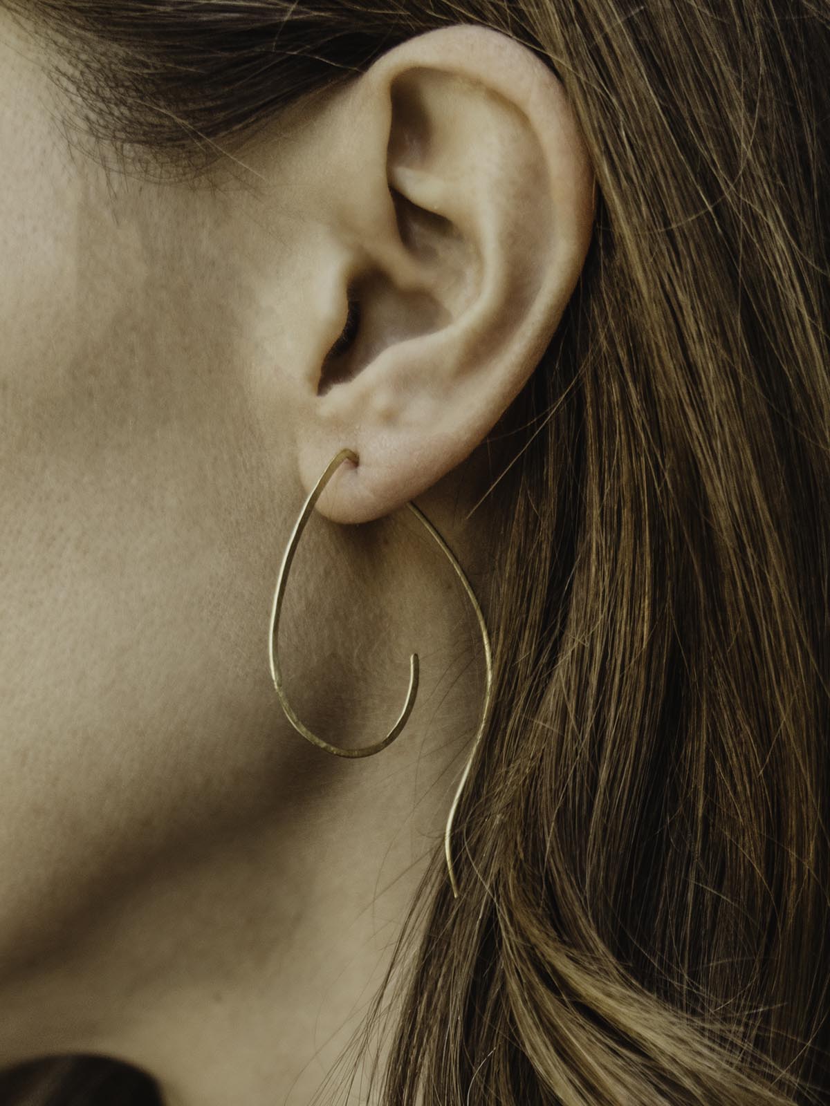 Close up of earring in models ear. Thin brass earring hangs from models ear.