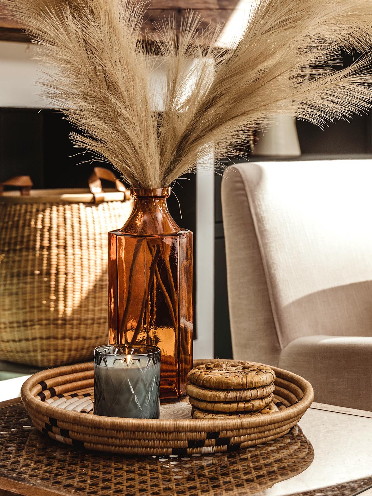 Stack of four banana leaf coasters inside a woven tray on a glass table with a blue candle and floral arrangement.