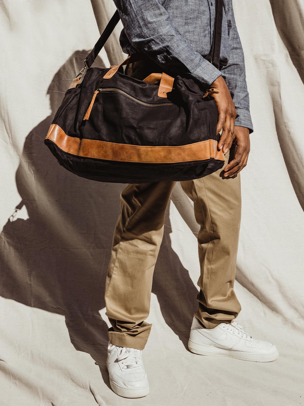 Male model wearing black weekender bag with brown leather accents. Bag is worn cross body on a cream cloth background.